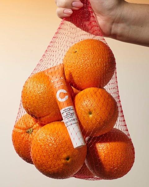 A high-contrast 4:5 image showing a Vitamin C skincare tube held inside a mesh bag filled with real oranges.